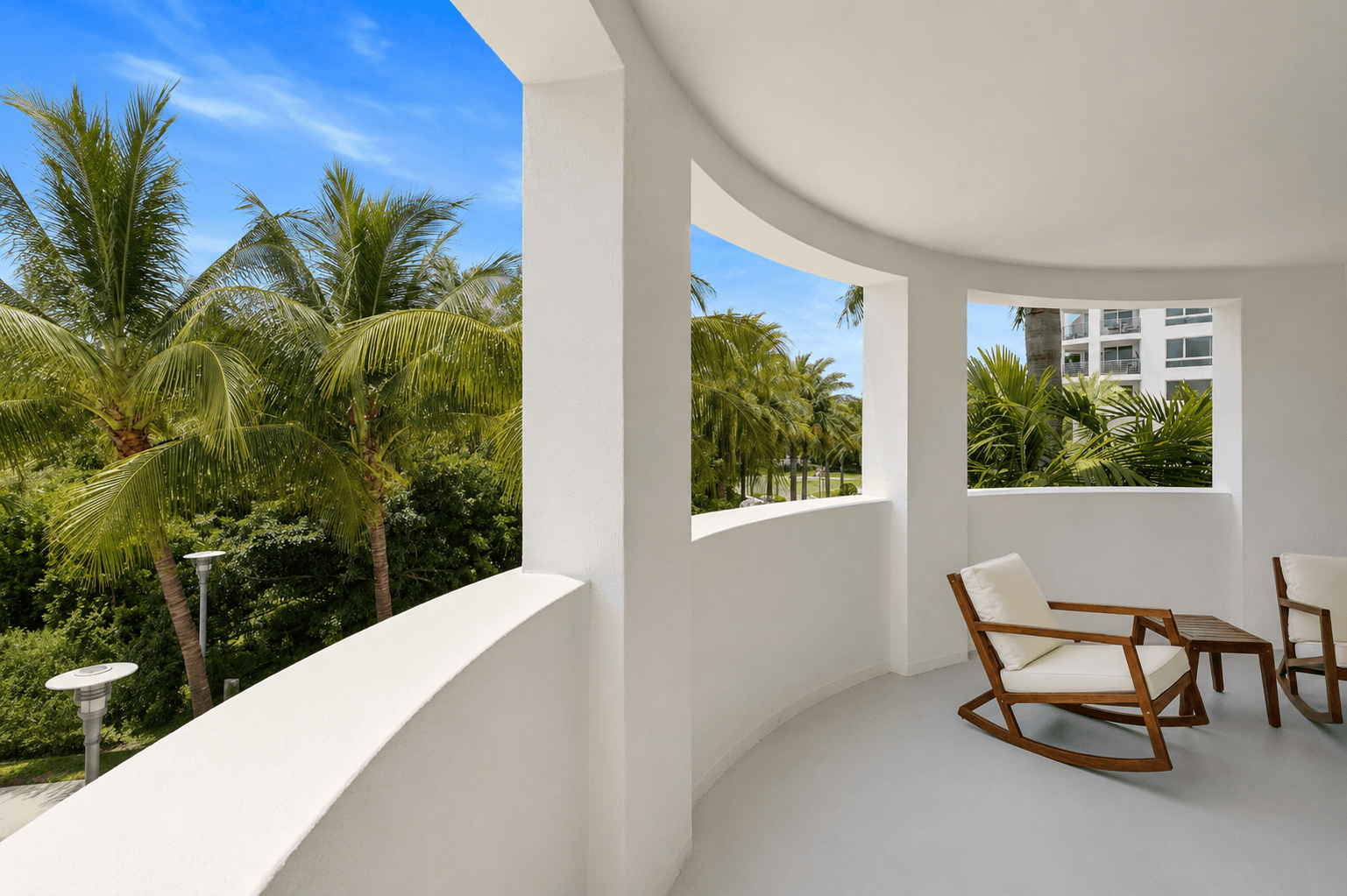 A white room with a wooden chair and table with a view of palm trees outside.